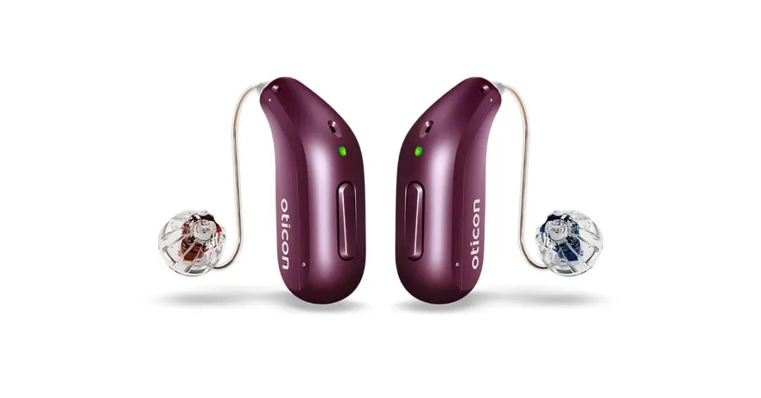 Two maroon Oticon hearing aids equipped with clear domes and attached thin wires are showcased side by side on a white background, featuring green indicator lights and sleek designs.